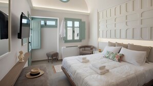 A bedroom with a bed, a chair, a bench, a TV, and a window with shutters. - Katerina Villas 11 12 13 & 3PriveJacuzzi (Santorini)