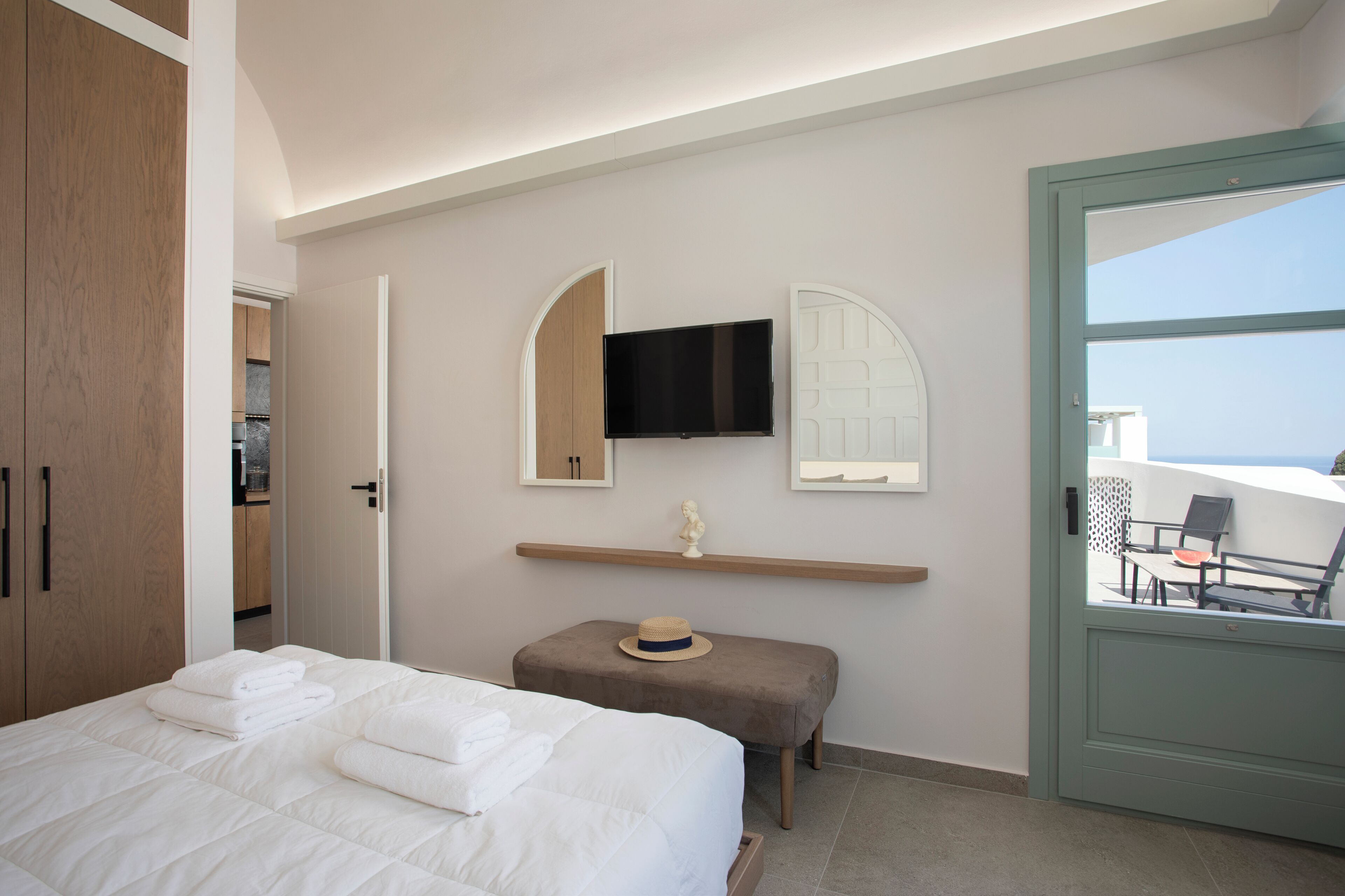A hotel room with a bed, a television mounted on the wall, a small table with a hat, and a view of the sea through a door.