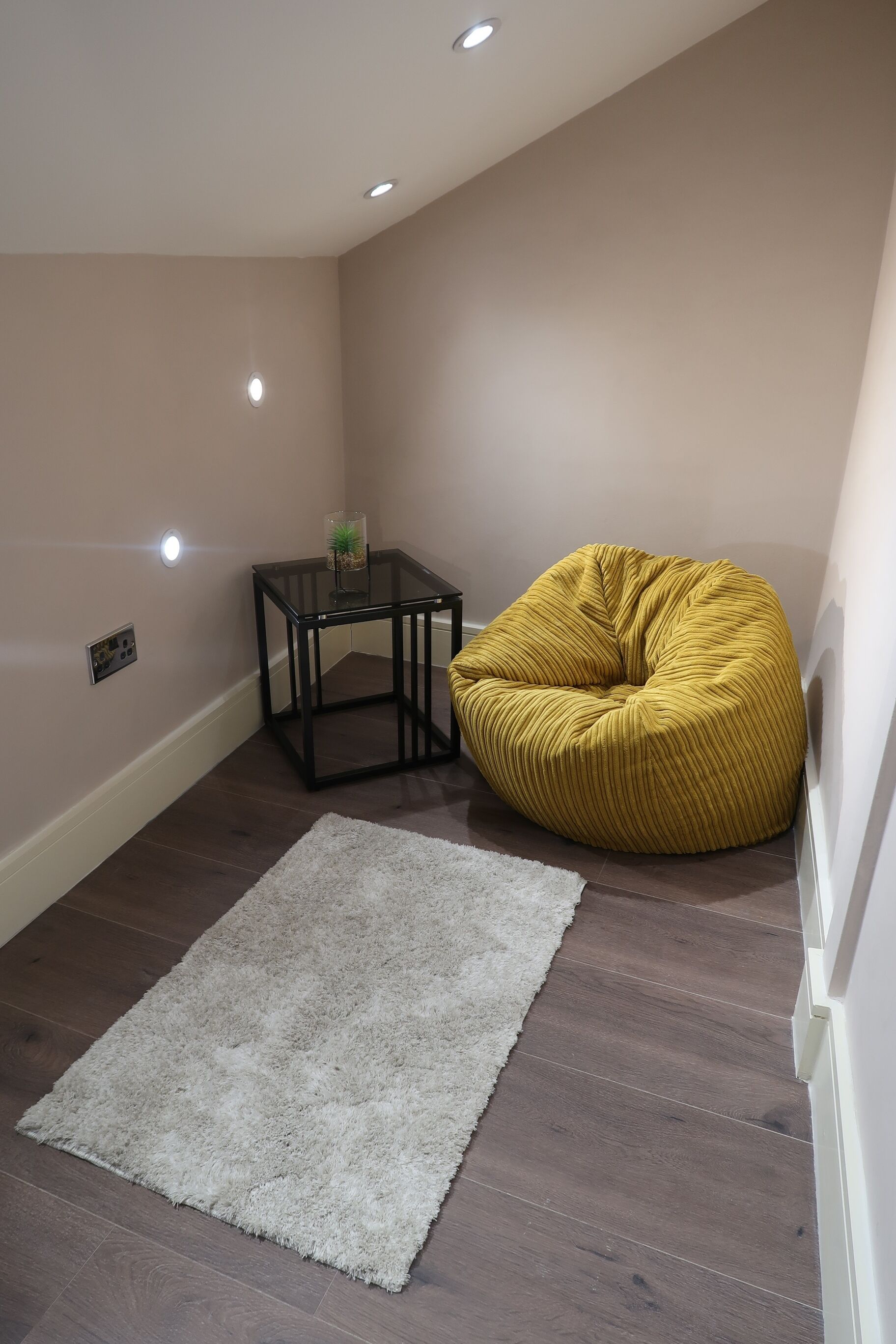 Ein Eckzimmer mit einem gelben Sitzsack, einem kleinen Glastisch mit einer Pflanze und einem weißen Teppich auf Holzdielen.