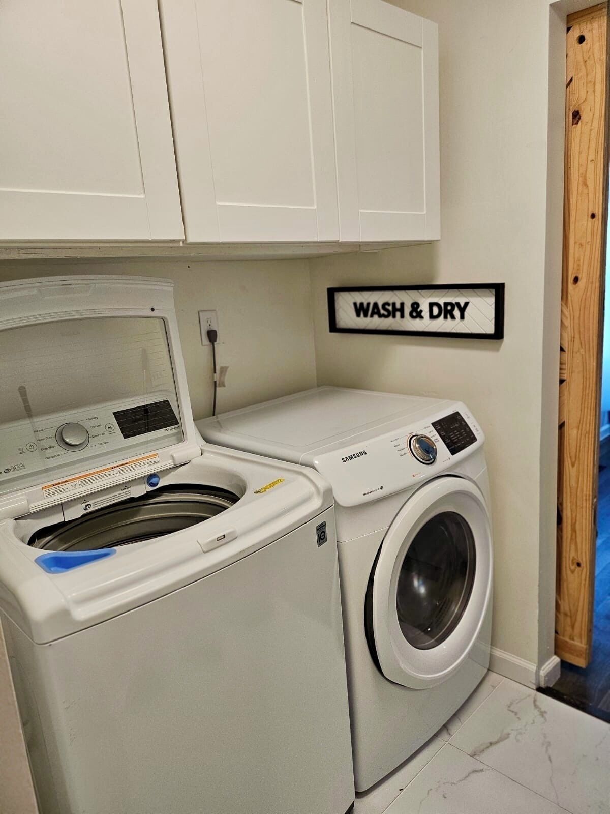A laundry room with a front-loading washer and dryer, a 'WASH & DRY' sign, and white cabinets above.