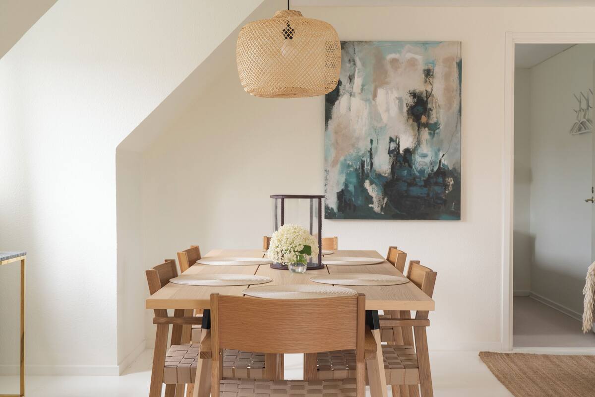 A dining area with a large wooden table, six chairs, a woven pendant light, and a modern abstract painting on the wall.