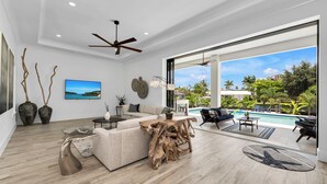 Ein modernes Wohnzimmer mit einer großen Couch, einem Couchtisch und einem Holzkunstwerk. Ein Fernseher ist an der Wand montiert, und es gibt einen Blick auf einen Pool und einen Außenbereich.
