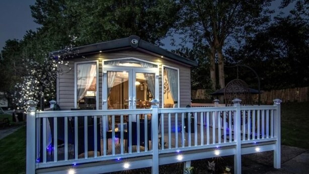 Et lille hus i et etage med en veranda og en terrasse, omgivet af træer og et hegn.