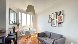 Sala de estar con sofá gris, suelo de madera, mesa de comedor con sillas, lámpara colgante y cuadros en la pared.