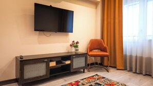 A modern living room with a flat-screen TV mounted on the wall, a dark wooden console table with shelves, a single orange chair, and a colorful rug on the floor. - GARDEN'S ROOM (Edirne)