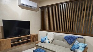A living room with a flat-screen TV on a wooden stand, a grey sofa with blue cushions, a small blue ottoman, and a window with vertical blinds. - Elegant 1 Bedroom - 1 bathroom Apartment (Tulum)