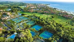 Vista aérea de un complejo turístico con varias canchas de tenis, una piscina y acceso a la playa.