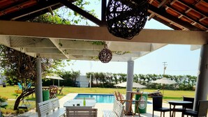 An outdoor patio with wooden tables and chairs, a pool in the background, and a covered area with hanging lights. - Pousada e Restaurante Lual do Parajuru (Beberibe)