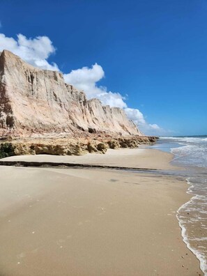Uma praia com areal arenoso e um penhasco rochoso.