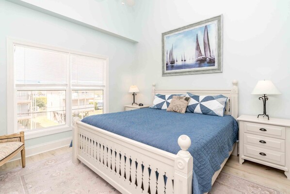 A bedroom with a bed, two bedside tables, a chair, a window with blinds, and a painting on the wall. - Meant To Be - Oceanfront 5 BR home with elevator! (Surf City)