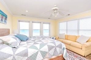 Quarto com cama, sofá, ventilador de teto e vista para o mar pelas janelas.