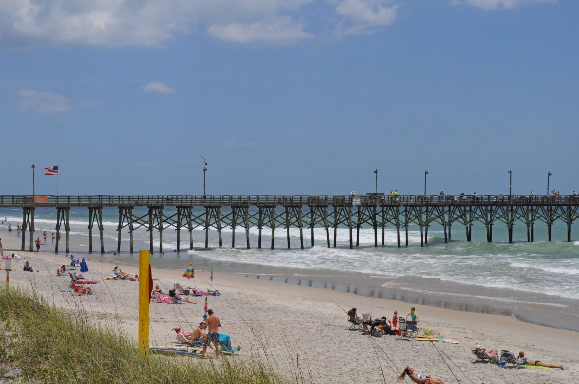 Ein Strand mit einem Steg, der sich ins Meer erstreckt, Menschen entspannen am Sand und eine Flagge auf dem Steg.
