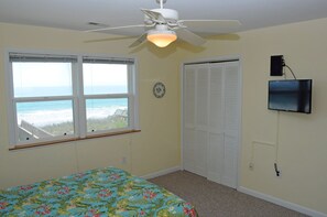 A bedroom with a bed, a ceiling fan, a window with a view of the beach, a closet, and a TV mounted on the wall.