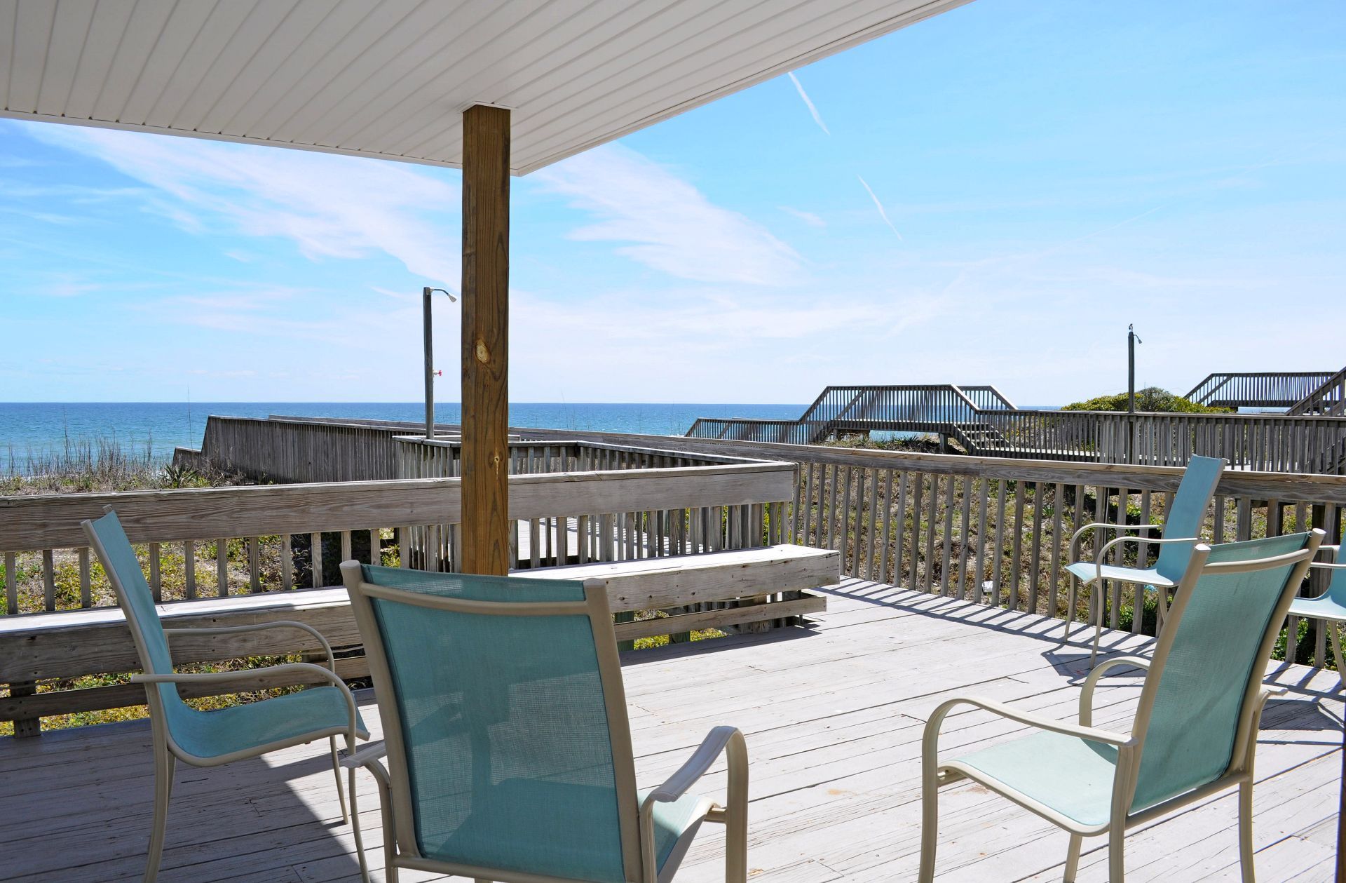 Une terrasse en bois, agrémentée de chaises et d’une table, qui offre une vue sur une plage avec une promenade en bois et un escalier menant à un niveau supérieur.