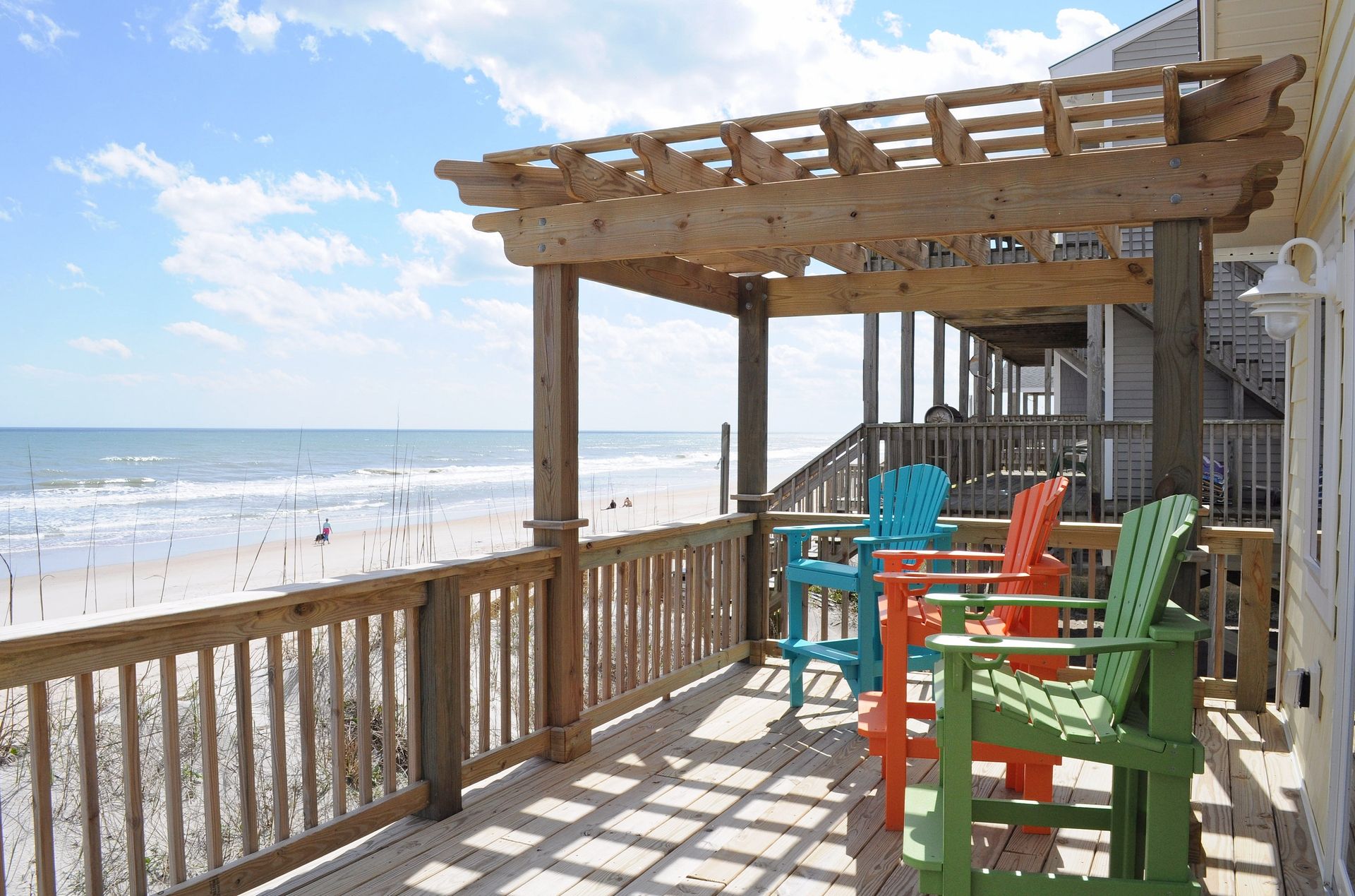 A wooden deck with a pergola, overlooking a beach with a view of the ocean and beachgoers.