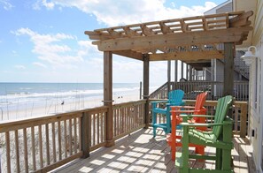 Eine Holzterrasse mit einer Pergola, mit Blick auf einen Strand und das Meer, sowie Badegäste.