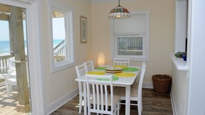 A dining area with a table set for four, white chairs, a hanging light fixture, and a view of the beach through a window. - Wholly Mackerel - Oceanfront, 2 Bedroom, Adorable Cottage in Surf City!  Dog (Surf City)