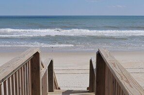 Ein hölzerner Steg führt zu einem Strand, im Hintergrund sind Wellen zu sehen.