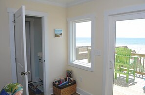 Ein Zimmer mit einer Tür, die auf einen Strand hinausführt, einem kleinen Fenster mit Blick aufs Meer und einem Korbwägelchen mit Gegenständen.