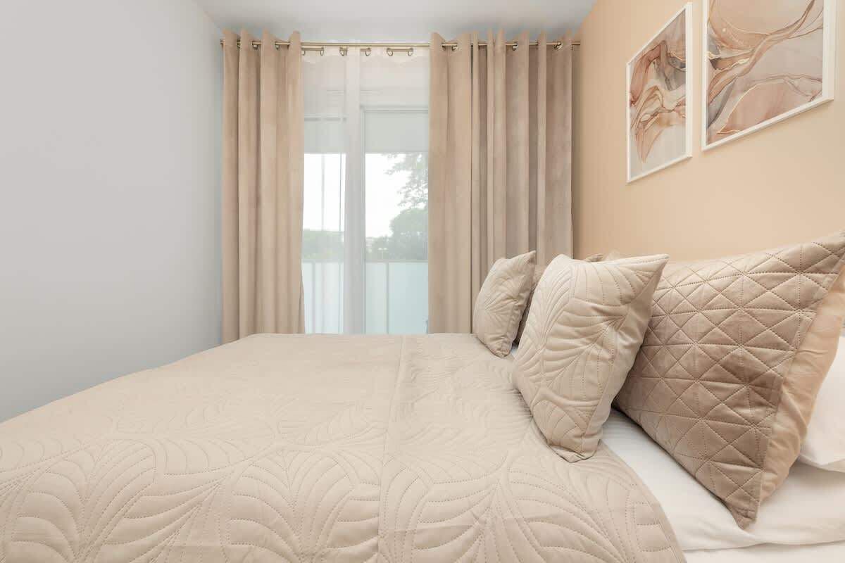 Ein Bett mit gesteppter Bettdecke, beigen Vorhängen und Blick ins Freie.