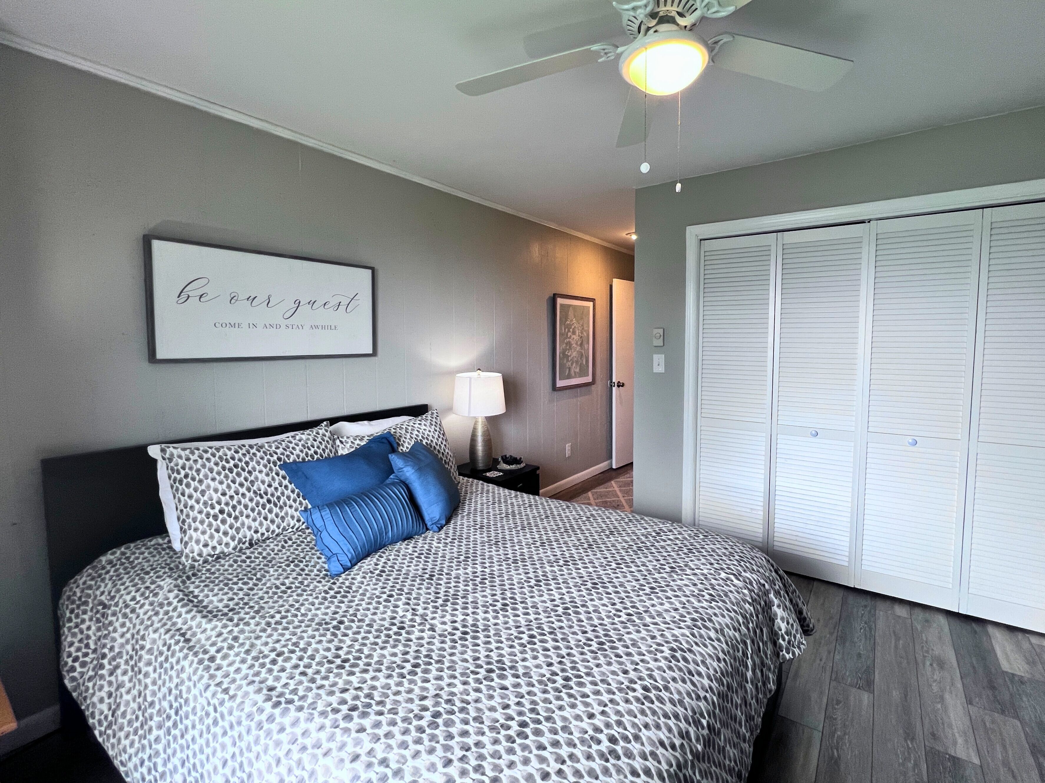 A bedroom with a bed, a ceiling fan, a framed sign, a nightstand, a lamp, and a wardrobe.
