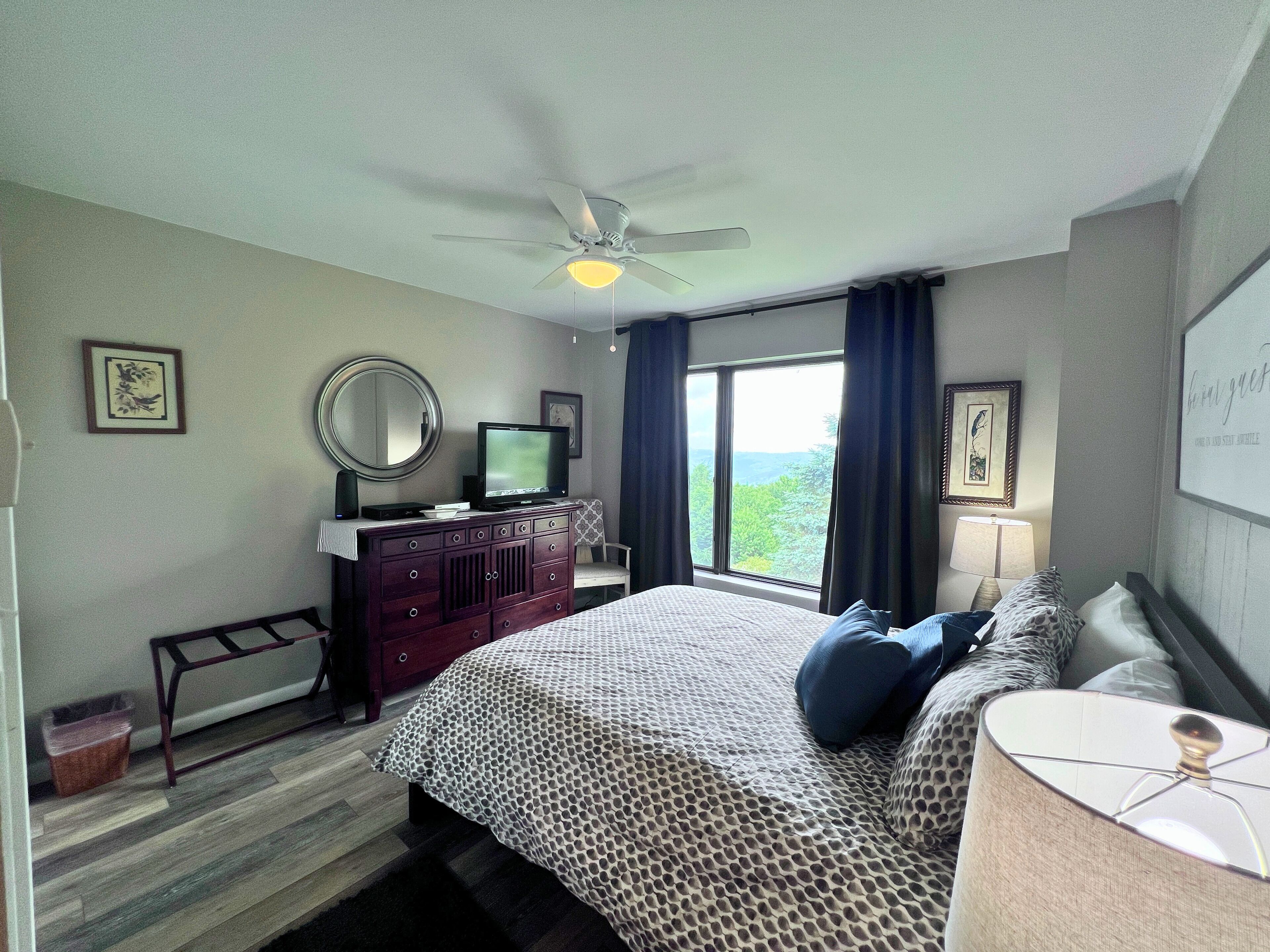 Une chambre à coucher comprenant un lit, une commode, un téléviseur, un ventilateur de plafond et une fenêtre donnant sur un beau paysage.
