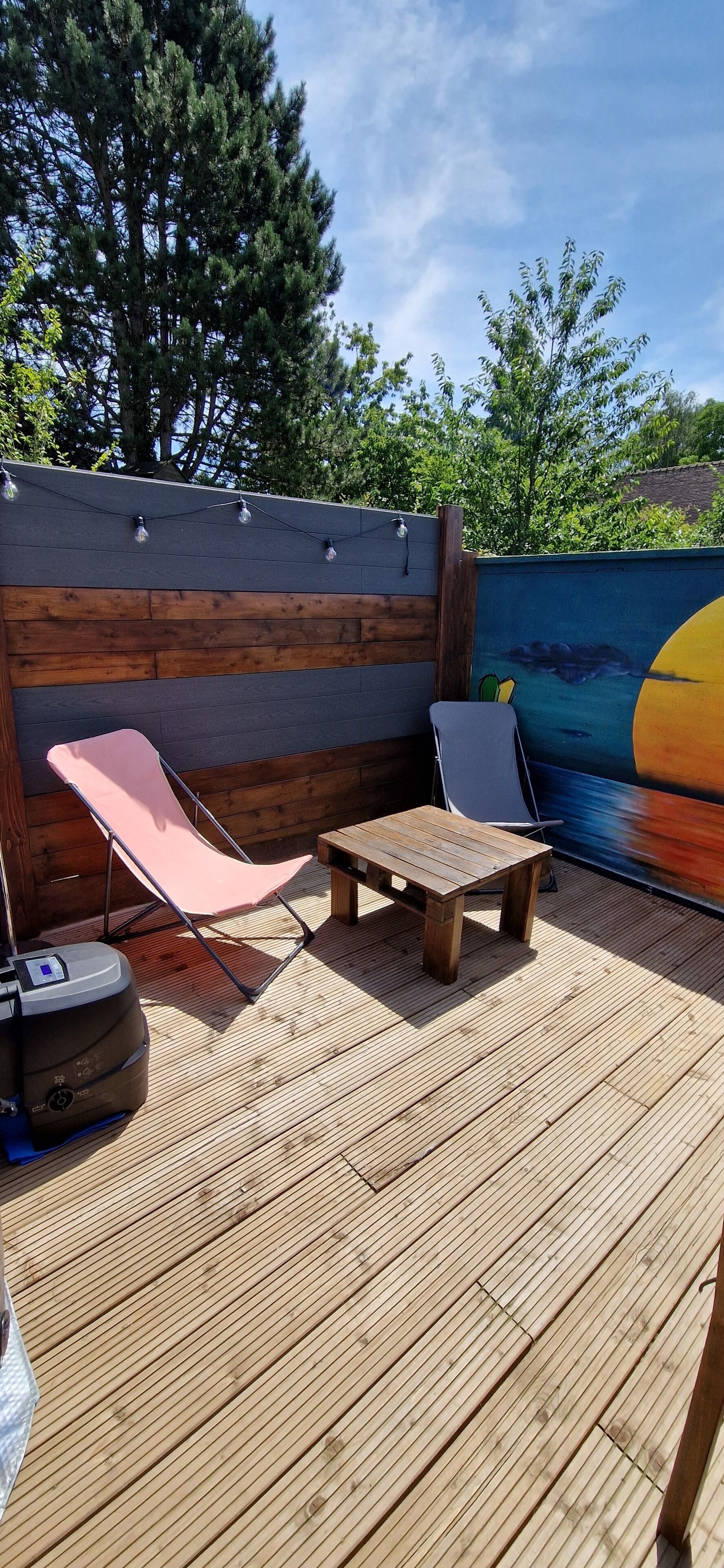 Una terraza de madera con dos sillones de descanso y una mesa, una parrilla para barbacoa y un mural en la pared.