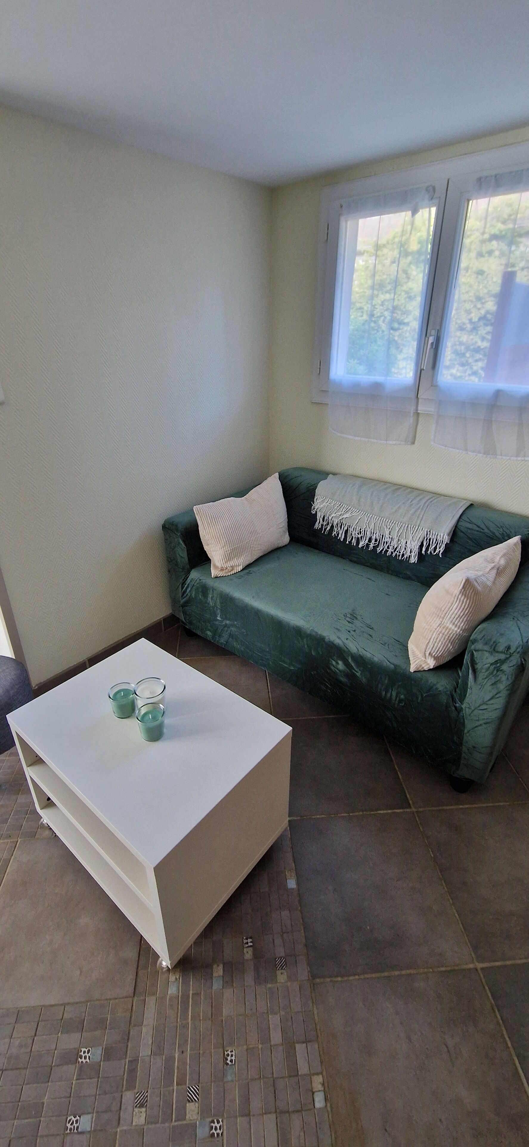 Una habitación con un sofá verde, una mesa de centro blanca y una ventana con cortinas de gasa.