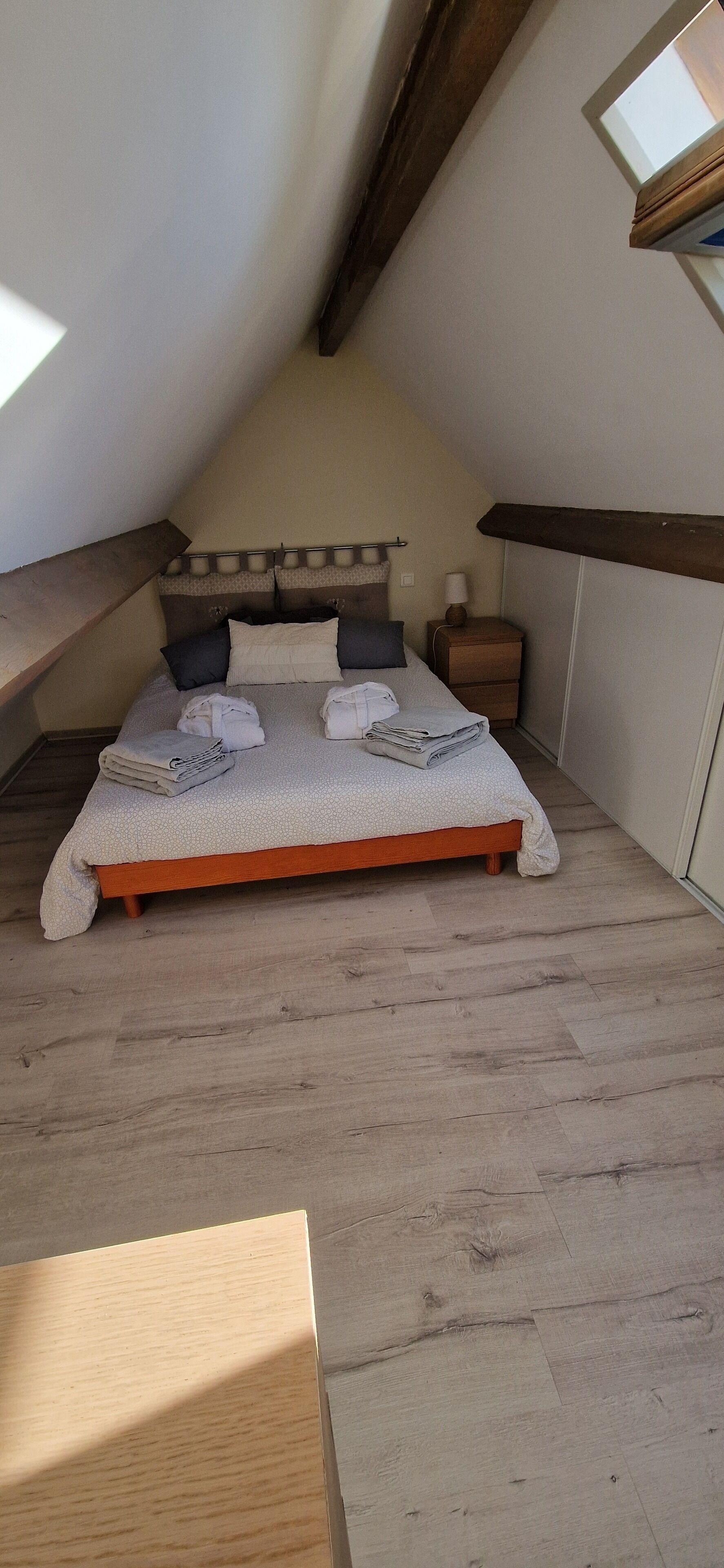 A bedroom with a sloped ceiling, a bed with a wooden frame, a nightstand with a lamp, and a small window.