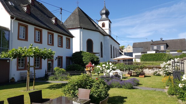 En traditionel bygning i europæisk stil med et spir, omgivet af en velplejet have med forskellige planter og blomster.