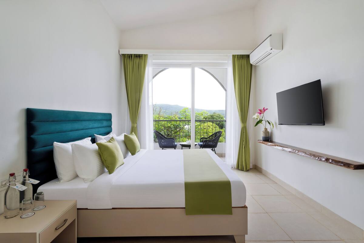 Habitación de hotel con una cama grande, televisión instalada en la pared y vistas a zonas verdes por la ventana.