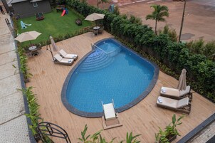 Una piscina con sillas de descanso y sombrillas, rodeada de follaje y un tobogán.