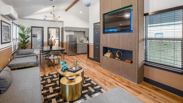 A modern living room with a sofa, coffee table, and a TV mounted on a wooden wall. There is a kitchen in the background with cabinets and a refrigerator. - HTX Ultra Chiic Suite #1 (Houston)