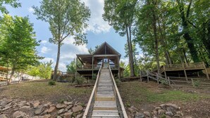 Un escalier en bois mène à un chalet doté d’un balcon, qui est entouré d’arbres et bordé par un terrain rocheux.