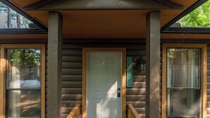 En stuga av trä med en vit ytterdörr, två fönster på var sin sida och en liten veranda med ett träräcke.