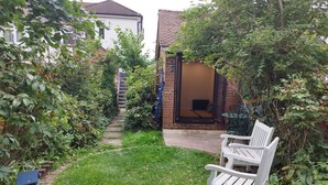 Un étroit sentier de jardin mène à une maison dont l’extérieur est en briques et qui possède une porte en bois. Un banc blanc se trouve au premier plan.