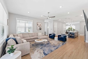 A spacious living area with a sofa, coffee table, and blue armchairs. A kitchen with a dining area is visible in the background.