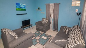 A living room with a blue wall, a flat-screen TV, a sofa, a chair, and a patterned rug. - Tao's Cozy Cottage! Piton View (Soufriere)