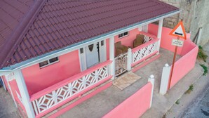 A pink house with a red roof, a porch with white railings, and a 'Reduce Speed Now' sign. - Tao's Cozy Cottage! Piton View (Soufriere)