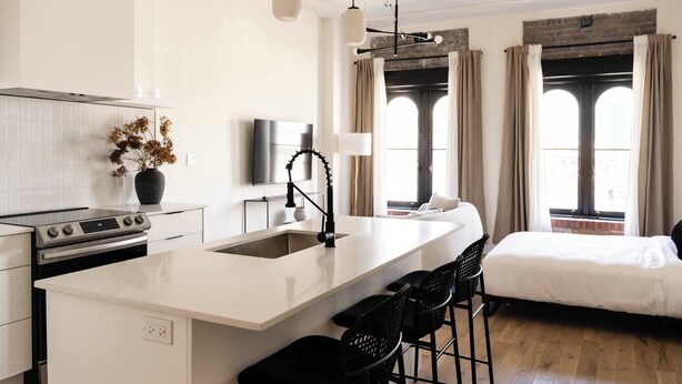 Une cuisine moderne avec un îlot central, un coin repas avec des tabourets de bar, un lit avec une tête de lit blanche et de grandes fenêtres avec des rideaux.