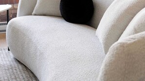 Un canapé blanc avec un coussin noir, des rideaux de couleur beige et une vue sur des immeubles par une fenêtre.