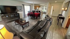 A living room with a TV, leather sofa, and a dining area with a red tablecloth. - Golfer’s Dream Home (Haines City)