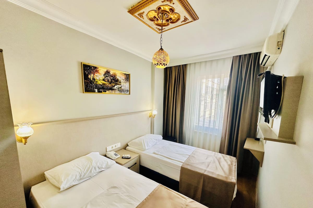 A hotel room with two beds, a painting on the wall, a ceiling light fixture, and a window with curtains.
