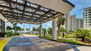 Un paseo techado con techo metálico geométrico, palmeras y un sendero pavimentado que conduce a un edificio.