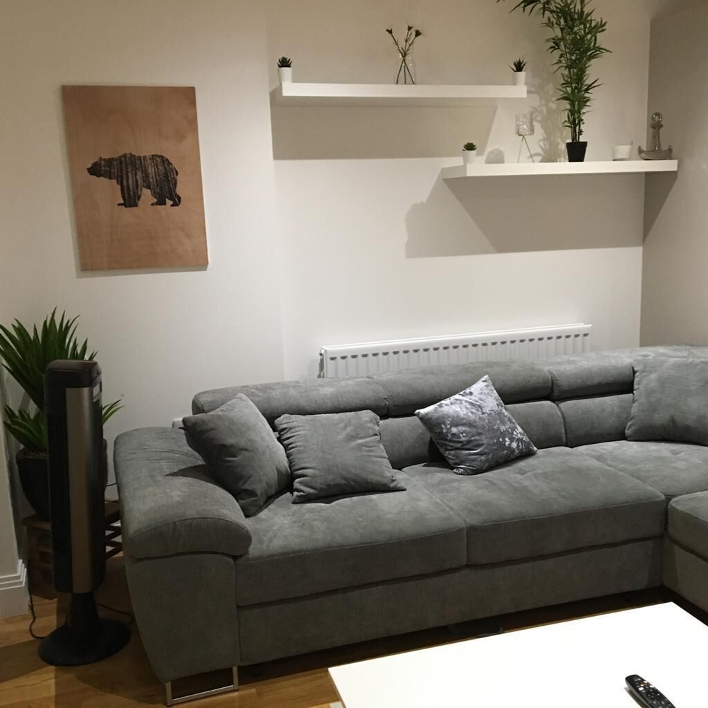Ein graues Ecksofa mit mehreren Kissen, ein Heizkörper darunter und ein Wandregal mit kleinen Pflanzen und Dekorationselementen.