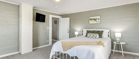 Un dormitorio con cama, ventilador de techo, televisión, una mesita de noche con lámpara y un cuadro en la pared.