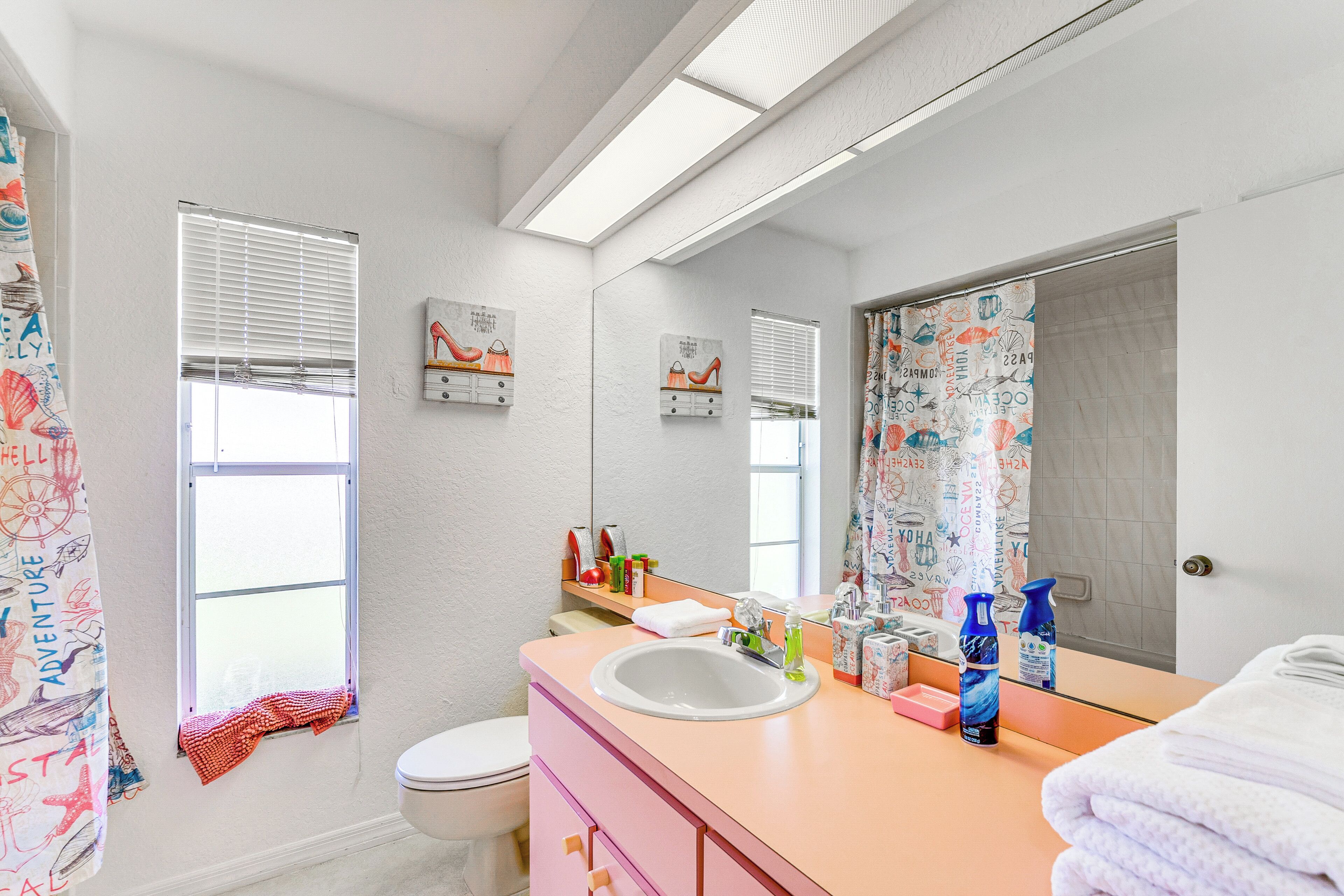 A bathroom with a white sink, pink vanity, and a large mirror. There are two windows with blinds, a shower curtain with a nautical theme, and a towel rack with folded towels.