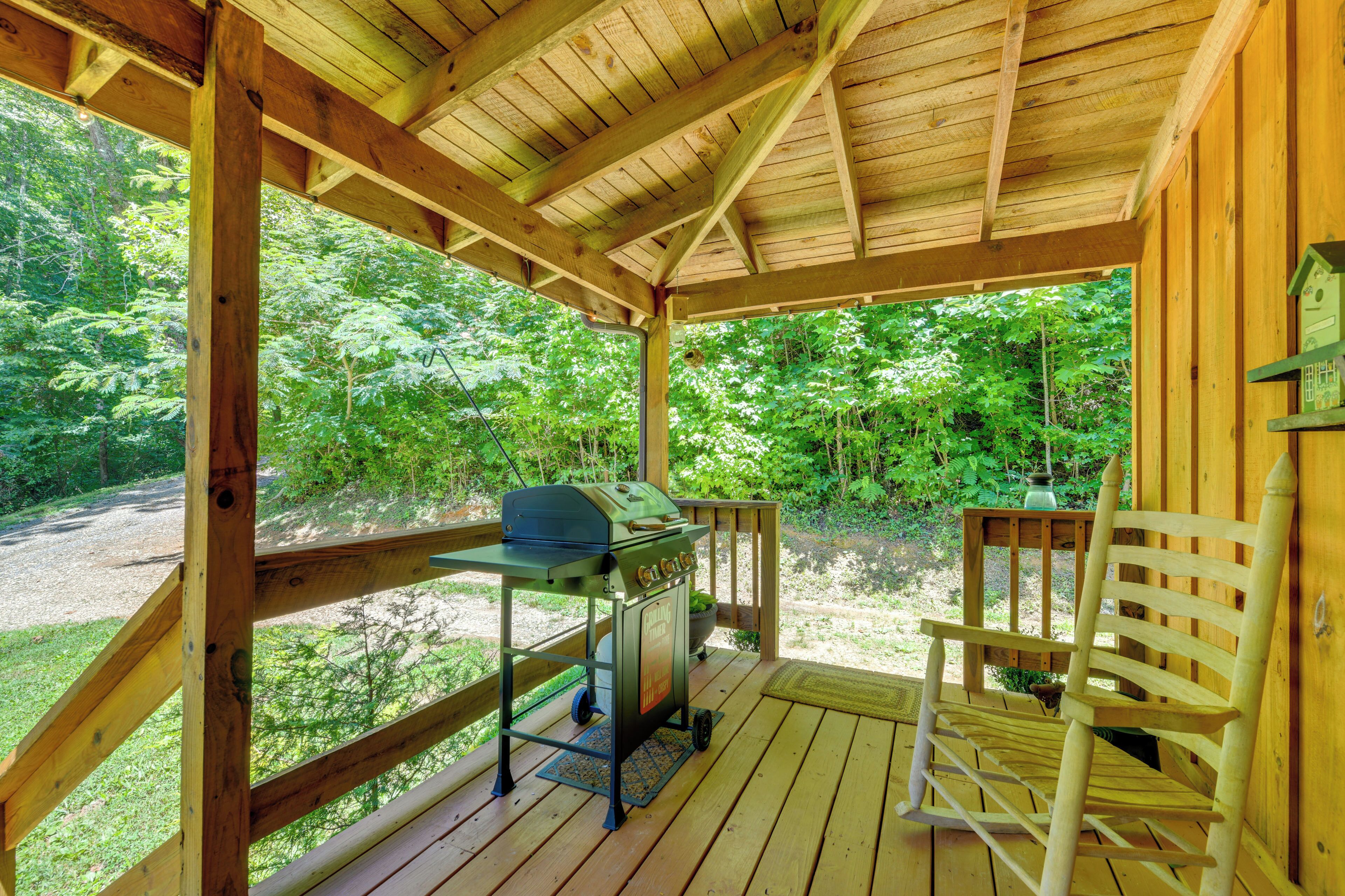 A wooden deck with a grill and a rocking chair, surrounded by lush greenery.
