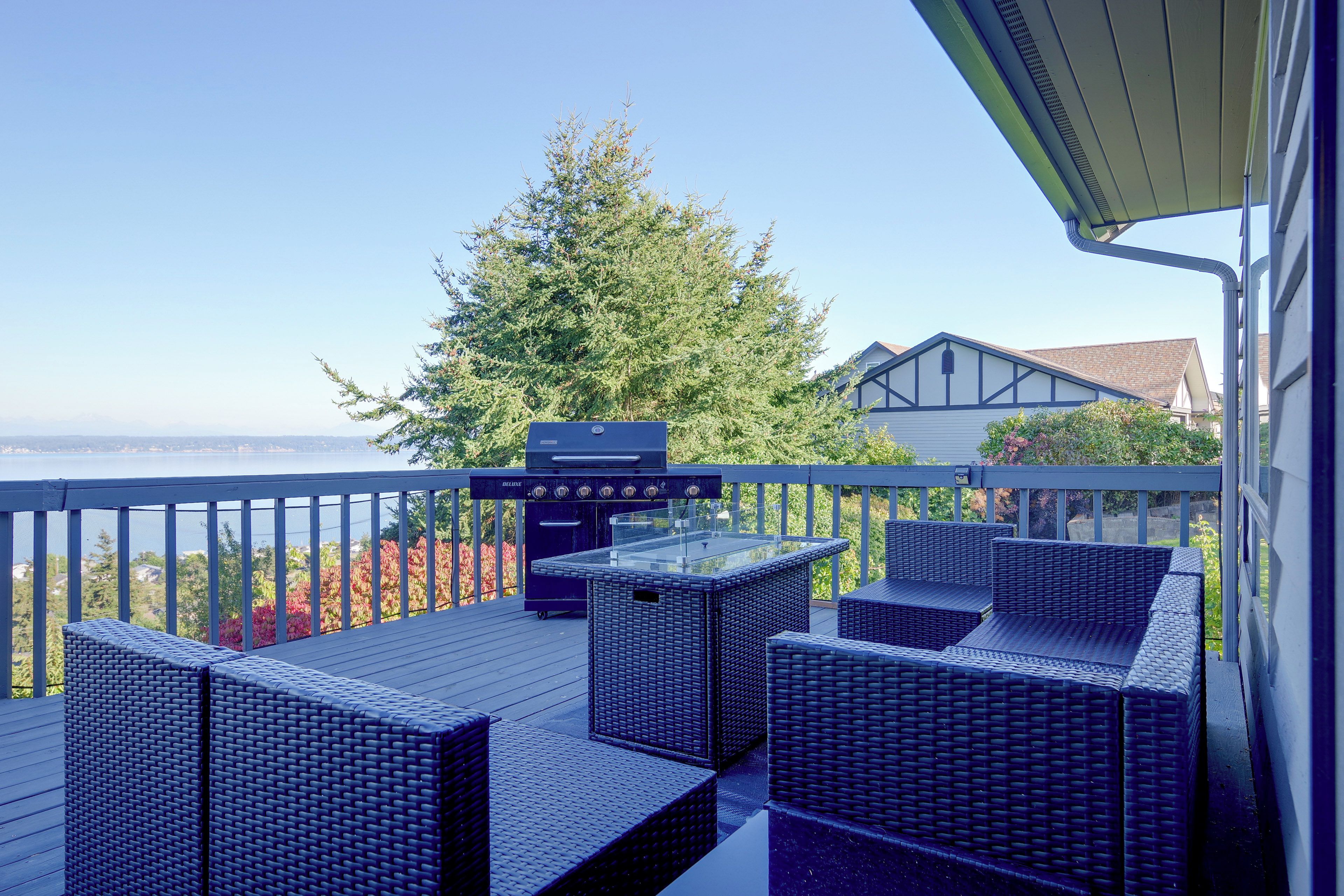 A deck with outdoor furniture and a grill, overlooking a body of water and a tree.