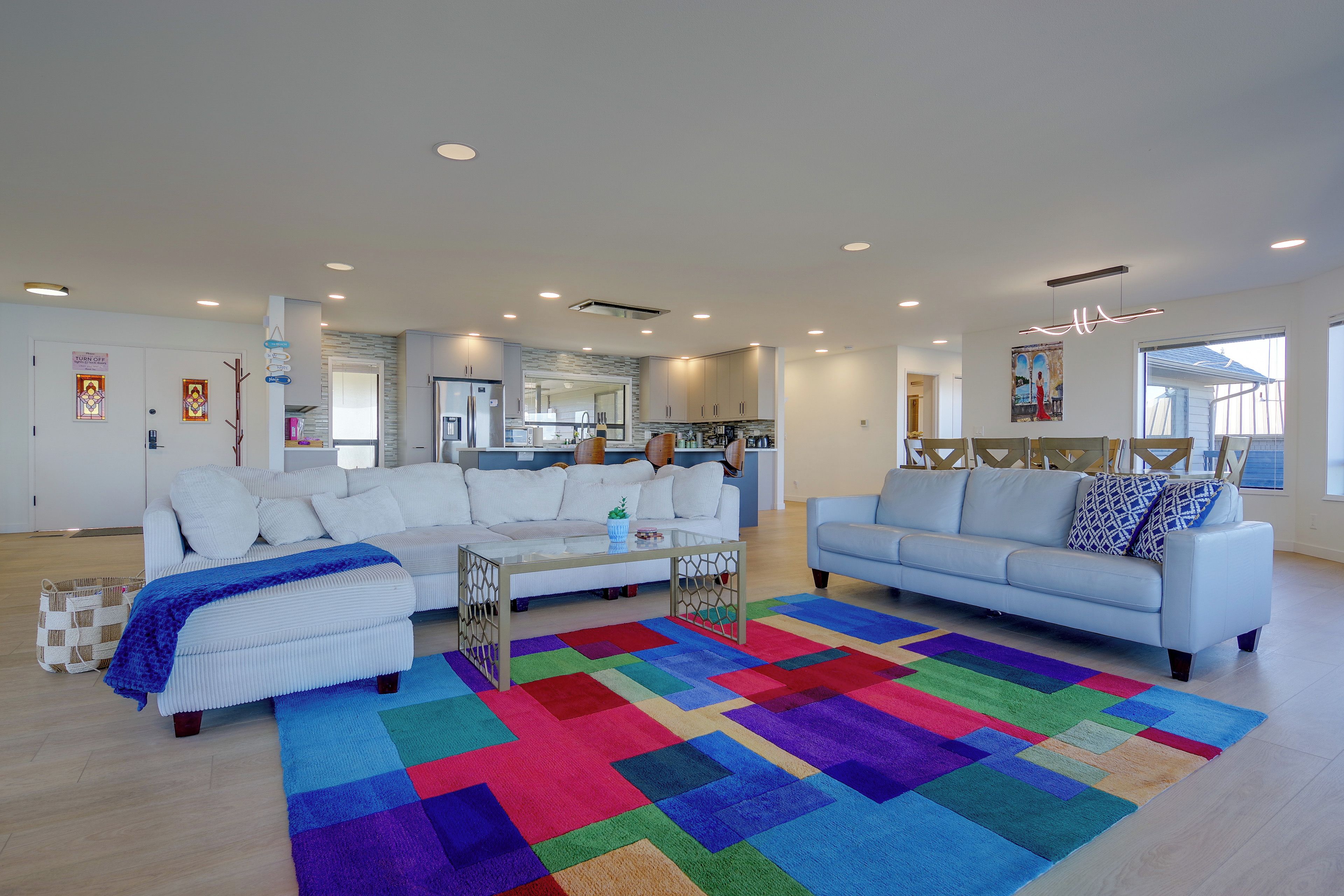 A spacious living room with a large colorful rug, two white sofas, a coffee table, and a kitchen in the background.